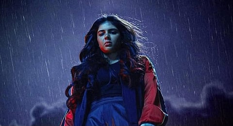 A woman with long wavy hair, wearing a dark outfit and red jacket, stands under heavy rain at night, lit dramatically against a cloudy sky.
