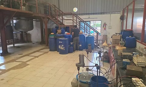 A cluttered industrial space with blue plastic barrels, chemical containers, and lab equipment. A man is standing near the barrels, facing away. The area includes metal stairs, large steel tanks, funnels, and glassware typically used in chemical processing. The scene suggests an illicit or makeshift chemical lab setup.
