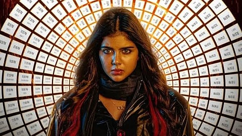 A woman with long dark hair streaked with red, wearing a black jacket and scarf, stands in front of a glowing tunnel-like backdrop covered in repeating text, staring intensely at the camera.