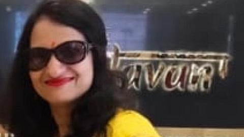 A woman wearing a bright yellow saree with a red embroidered blouse, accessorized with dark sunglasses and a small red bindi on her forehead. She has long black hair, styled loosely, and is smiling. She is holding a smartphone in her hand, with a mehndi design visible on her wrist and hand. The background shows part of a signboard and marble wall decor.