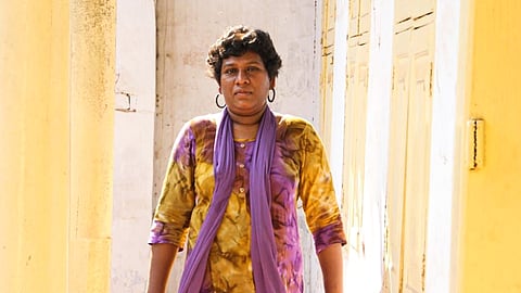 Bindhu Ammini against a yellow backdrop, facing the screen and looking at the viewer