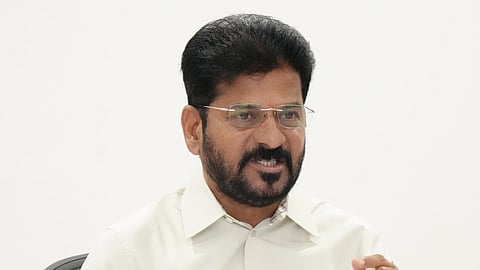 A man with short black hair, beard, and glasses is seated at a desk, holding papers in one hand and gesturing with the other. He is wearing a light-colored shirt and appears to be speaking, possibly in a meeting or discussion. Documents, a phone stand, and electronic devices are on the desk.