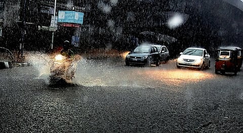 Heavy rainfall in Hyderabad floods a city road, with a scooter, cars, and an auto-rickshaw navigating through waterlogged streets as headlights reflect off the wet surface.