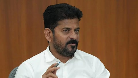 CM Revanth Reddy with short black hair and a beard is sitting at a table, wearing a white shirt. He is holding a pen in his right hand and appears to be speaking or making a point. A microphone is placed in front of him, along with a notebook, water bottle, and a holder with various pens. The background is a wooden panel.
