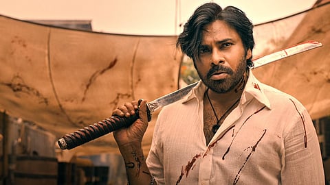 Pawan Kalyan, looking rugged with messy hair and a blood-stained white shirt holds a large sword resting on his shoulder. His intense expression, smudges of blood on his face and clothes, and the dramatic backdrop suggest a fierce, action-packed scene.