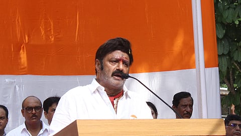 A man wearing a white shirt is speaking at a podium with a microphone during a public event. He has a red tilak on his forehead and is standing in front of an orange and white backdrop. Several people in white shirts are standing behind him. The podium has the name "BASAVATARAKAM" on it.