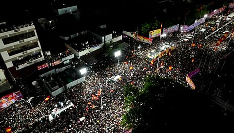 A massive crowd gathers tightly packed on the streets of Karur at night for a TVK political rally, with floodlights, buses, flags, and banners visible, and people filling rooftops and surrounding buildings.