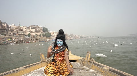 A low-angle, wide shot shows a young child in the attire of the Hindu deity Lord Shiva sitting on the bow of a wooden boat on a river, likely the Ganges (Ganga), in Varanasi, India.
The child, facing the camera, has their face painted blue with white markings (similar to Shiva's third eye), wears a high black wig styled into a bun, and has a garland of plastic black "snakes" around their neck, one of which rests on their shoulder. They are wearing a brightly patterned, orange and black tiger-print cloth, suggesting the traditional depiction of Shiva's tiger skin. The child is raising their right hand in a wave.
In the background, the river is wide and murky, with many white seabirds flying or floating on the water. On the left bank, the dense, ancient stone and stucco buildings of a ghat in Varanasi rise up. The sky is a pale, hazy white. The foreground of the wooden boat is dominated by the child and the worn, weathered wood of the vessel.