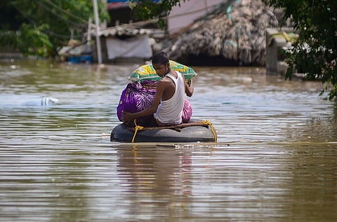 A person in a white tank top and dark shorts sits on an inflatable rubber inner tube, likely makeshift raft, in a large expanse of brown floodwater. They are holding two bundles: one wrapped in a purple cloth and the other in a green and yellow striped sack, possibly carrying belongings. The water is still but reflective. In the background, partially submerged structures and vegetation are visible, suggesting the area is a flooded residential or rural location.
