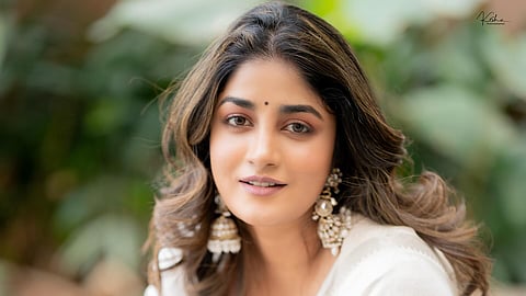 A head-and-shoulders, medium-close-up shot of actress Dimple Hayathi seated outdoors, looking directly at the camera with a gentle expression.
She is wearing a white saree and a matching blouse with a V-neckline. Her long, dark brown hair is styled in loose waves. She's accessorized with large, ornate white and gold jhumka earrings and a small black bindi. Her jewelry also includes several bangles on her right wrist, some of which are green and white, and a prominent diamond ring on her left index finger. The background is a softly blurred outdoor setting dominated by green foliage.