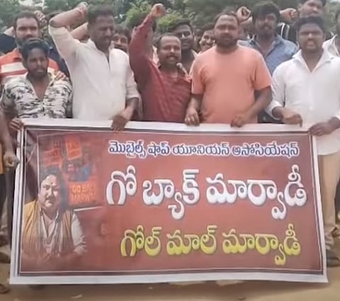 A group of men is holding a large red and white banner in what appears to be an outdoor protest. The men are mostly raising their fists in the air. The banner features text in Telugu, which includes:
'మొబైల్స్ షాప్ యూనియన్ అసోసియేషన్' (Mobiles Shop Union Association)
'గో బ్యాక్ మార్వాడీ' (Go Back Marwadi)
'గోల్ మాల్ మార్వాడీ' (Golmaal Marwadi, which can be interpreted as 'Fraud/Deceptive Marwadi').
There is also a picture of a man on the left side of the banner with small text that includes 'GO BACK MARWADI' in English. The image captures a demonstration against Marwadi traders by a Mobile Shops Union Association.