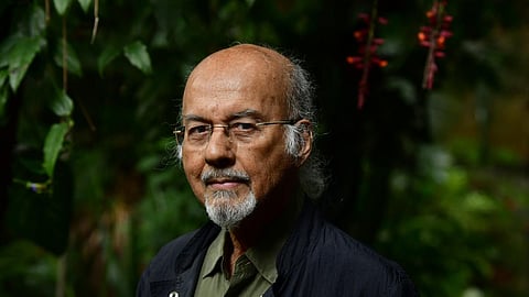 A headshot of an elderly man with a bald head and a gray goatee, wearing glasses and a dark jacket over a green shirt. He looks directly at the camera with a neutral expression. The background is blurred but appears to be a lush, green outdoor setting with some hanging red flowers visible at the top right.
