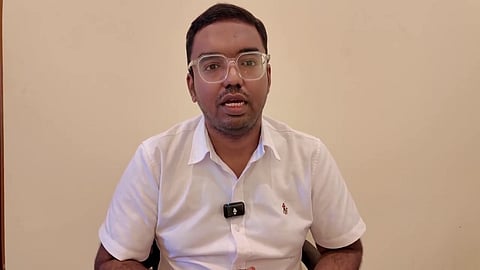 A man wearing a white collared shirt and modern glasses is speaking directly to the camera. He has a small, black wireless microphone clipped to his shirt.