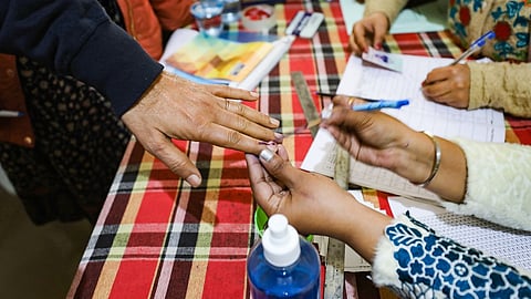 Close-up image of a person's hand receiving an indelible ink mark on their finger at a voting booth.
A pair of hands, with a light-tan complexion and wearing a white cuff, holds the thumb and index finger of a voter's hand, which is stretched across a checkered red, black, and white table. The voter's hand has a wedding band on the ring finger and is being marked with purple/indelible ink on the fingertip, a common practice to indicate that a person has voted.
In the background, on the right side, another person is writing on a sheet of paper. Several items are scattered across the table, including a clear plastic bottle with "AQUA" written on a blue label, a ruler, pens, and a smaller bottle of blue liquid, possibly sanitizer. The setting appears to be an indoor polling station.