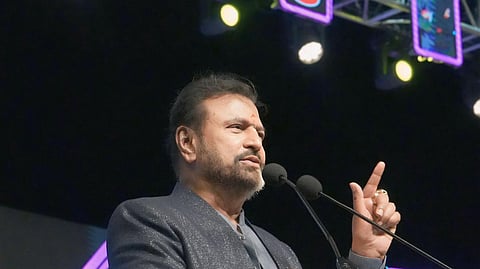 A medium-shot photograph of Mohan Babu speaking at a podium under a stage setup for Mohan Babu University (MBU). He is a middle-aged man with a light beard and mustache, wearing a dark, textured suit jacket over a dark shirt, and has a small red mark (tilak) on his forehead. He is gesturing with his right hand, pointing his index finger upwards, and looking slightly to his left. The MBU logo and university name are prominent on a large banner behind him, illuminated in red and white light, with a smaller vertical MBU logo and a display screen showing a picture of the actor/founder on the right.