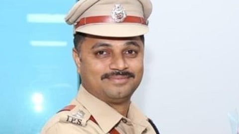 A man wearing a tan police uniform with badges and insignia of the Telangana Police, standing indoors and smiling slightly while holding a bouquet.