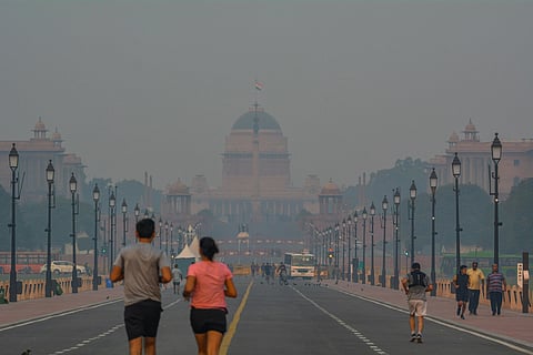 People jogging and walking along a smog-covered Rajpath in New Delhi, with the Rashtrapati Bhavan barely visible through thick haze caused by air pollution.