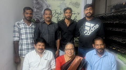 A posed indoor group photo of seven people—three older adults seated in the front row and four younger men standing behind them.
The front row features a woman in a red and black saree flanked by two men in collared shirts (white and light blue). The back row features four men, including one in a plaid shirt and another in a black Adidas hoodie. Everyone in the group is smiling. The background includes a decorative wall design.