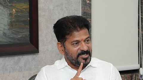 Revanth Reddy sitting at an office desk, wearing a white shirt, and emphatically speaking while raising his index finger.