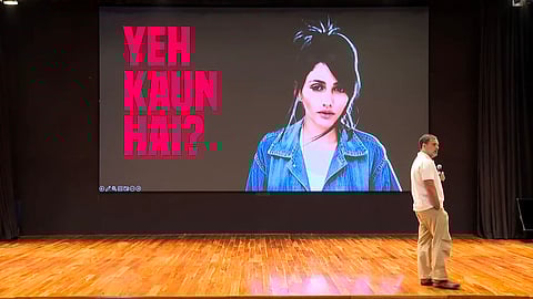Rahul Gandhi is standing on a stage with a microphone, facing a large screen that displays an image of a woman in a denim jacket next to bold pink Hindi text reading “Yeh Kaun Hai?” (meaning “Who is this?”). The setting appears to be an indoor auditorium with wooden flooring.