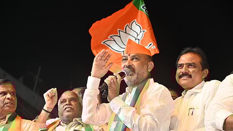 A group of men at a political rally, one man speaking into a microphone while wearing an orange cap and scarf with the BJP (Bharatiya Janata Party) logo. A large BJP flag with the lotus symbol is visible behind them, and the men appear to be addressing a crowd at night.