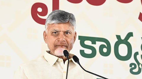 A close-up photograph of N. Chandrababu Naidu, an Indian politician, standing at a clear podium and speaking into two microphones. He is wearing a light cream or off-white collared shirt, and has grey hair and a light beard. He is holding a piece of paper in his right hand. In the background, there is a stage banner with Telugu script and the partially visible English text "FIBER" and "EMBER".