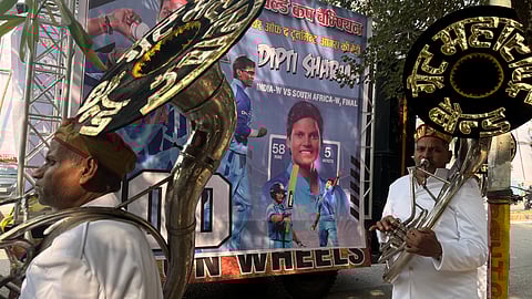 Two band musicians dressed in white uniforms and red-gold turbans stand on a street playing brass instruments during a celebration. Behind them is a large poster of cricketer Deepti Sharma, highlighting her World Cup performance, with images of her batting and bowling. Trees and decorations surround the scene, indicating preparations for a festive welcome.