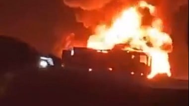 A low-quality, dark photograph showing a massive fire engulfing what appears to be a large vehicle, possibly a tanker truck, on a highway at night. Intense bright orange and yellow flames and thick smoke rise high into the air, illuminating the scene. A second vehicle, partially visible on the left, has its headlights on, providing some contrast. The surrounding area is very dark.