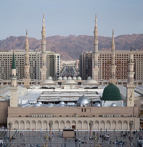 An eye-level, wide shot captures the immense scale of Al-Masjid an-Nabawi (The Prophet's Mosque) in Medina, Saudi Arabia, against a backdrop of modern high-rise buildings and distant reddish-brown mountains.
In the foreground is the vast, open marble courtyard filled with numerous people. The main structure of the mosque is a complex of beige stone walls featuring numerous arched openings and several large, gray domes.
The most distinctive feature is the Green Dome (Qubbat an-Nabi), located over the resting place of the Prophet Muhammad, which stands prominently on the right side of the mosque complex. To the left of the Green Dome, another taller section of the building features a single slender minaret topped with a green finial.
Rising from the mosque complex are several very tall, pale beige minarets with multiple balconies and pointed tops, dominating the skyline. Behind the mosque, a dense collection of multi-story modern hotels and residential buildings spans the background, with a wide, bustling avenue cutting through the city center. The arid mountains form the final layer of the backdrop.
