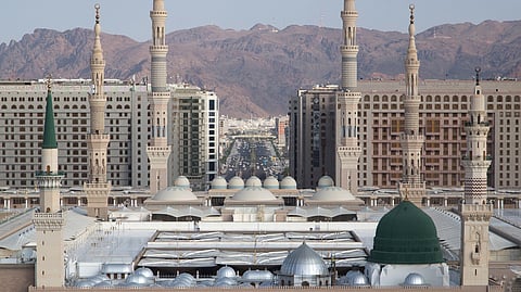 An eye-level, wide shot captures the immense scale of Al-Masjid an-Nabawi (The Prophet's Mosque) in Medina, Saudi Arabia, against a backdrop of modern high-rise buildings and distant reddish-brown mountains.
In the foreground is the vast, open marble courtyard filled with numerous people. The main structure of the mosque is a complex of beige stone walls featuring numerous arched openings and several large, gray domes.
The most distinctive feature is the Green Dome (Qubbat an-Nabi), located over the resting place of the Prophet Muhammad, which stands prominently on the right side of the mosque complex. To the left of the Green Dome, another taller section of the building features a single slender minaret topped with a green finial.
Rising from the mosque complex are several very tall, pale beige minarets with multiple balconies and pointed tops, dominating the skyline. Behind the mosque, a dense collection of multi-story modern hotels and residential buildings spans the background, with a wide, bustling avenue cutting through the city center. The arid mountains form the final layer of the backdrop.