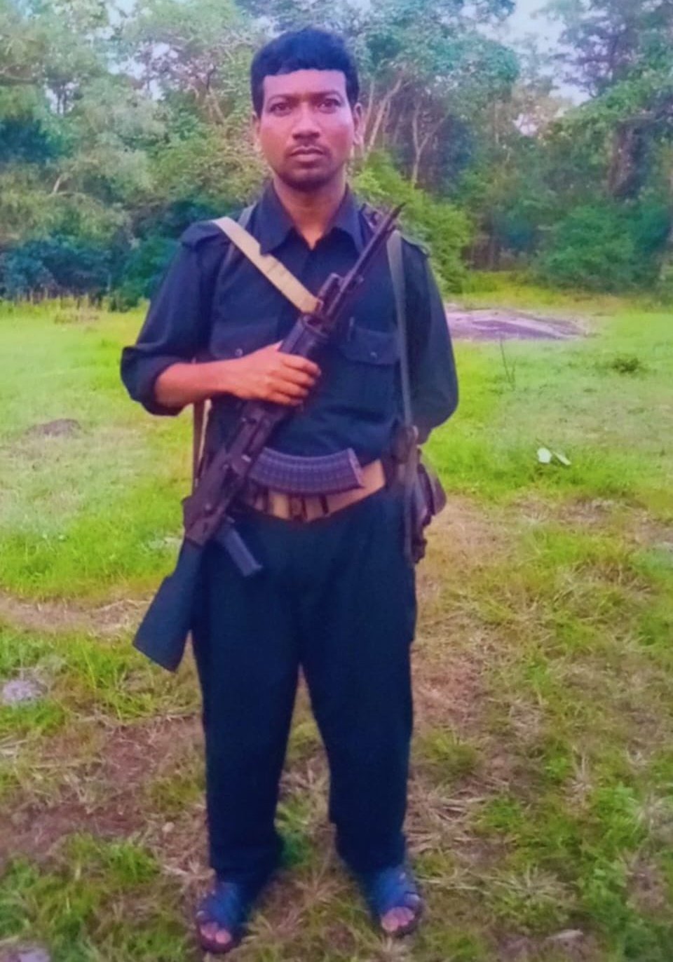 A medium-shot photograph of a man standing outdoors in a grassy, forested area. He is wearing dark blue or black clothing, appearing to be a button-down shirt and trousers. He is holding an assault rifle, likely an AK-47 variant, diagonally across his chest with a strap over his shoulder. He has an ammunition belt around his waist and is wearing sandals on his feet. The background consists of green grass and dense forest foliage with some rock formations visible in the distance.
