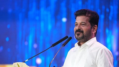 A portrait photo of Revanth Reddy, an Indian politician, addressing an audience. He is a middle-aged man with a beard and dark hair, wearing a crisp white shirt and standing at a podium with two microphones. The background is a vibrant, blurred blue light with a bright stage setup, and the foreground features an arrangement of white and blue flowers, including what appear to be Calla Lilies and Orchids.