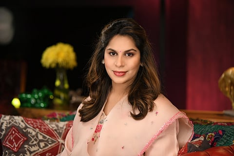 A medium close-up photo of Upasana Konidela, a woman with warm brown hair and a pink bindi, looking directly at the camera with a gentle smile. She is wearing a light pink traditional Indian outfit, possibly a saree or salwar kameez, with subtle pink embroidery along the border of her dupatta. She is seated on a richly patterned couch, with a blurred, dark background featuring a vibrant yellow floral arrangement on the left.