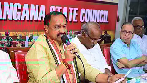 A political leader, Mahesh Kumar Goud, wearing a light yellow kurta and a traditional scarf with black and red patterns, is seated at a table and speaking into a microphone. He is holding a piece of paper in his left hand. Behind him, a banner with red and white text partially reads "TELANGANA STATE COUNCIL". Several older men are seated next to and behind him, listening. The man immediately to his right is wearing a white shirt and looking down, and the man further right is wearing a light blue shirt and glasses and looking forward.