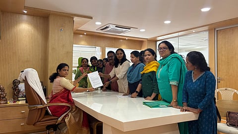A group of approximately ten women, appearing to be journalists, are standing around a large, white conference table in a modern office. One woman, seated in a chair on the left, is wearing a red and cream saree and is holding a piece of paper, seemingly reviewing it or accepting it from the group. The other women are standing on the opposite side of the table, looking towards the seated woman. One woman in the center, wearing a light beige outfit, is extending her hand towards the paper. The office walls are paneled with light wood, and a small shrine or idol is visible on a cabinet to the far left. The scene suggests a formal meeting or the submission of a document, possibly a complaint, given the filename.