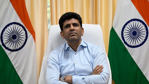 A head-on, medium shot of a man identified as Pemmasani Chandrasekhar sitting in a white chair behind a dark wooden desk.
He is wearing a light blue, long-sleeved collared shirt and a silver wristwatch.
He is looking directly at the camera with a serious expression, his arms crossed on the desk in front of him.
Behind him, on the left and right, are two Indian flags (Tiranga) displayed vertically.
The background features light yellow/cream curtains on either side of a central wall.
A piece of paper and a pen are visible on the desk.