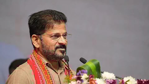 A close-up shot of an Indian politician, Revanth Reddy, speaking at a podium. He is a middle-aged man with a neatly trimmed beard, wearing glasses, and a light brown or beige shirt. A red and white patterned shawl/scarf is draped around his neck. In the foreground, there is a large arrangement of colorful flowers, including white, purple, and yellow blossoms. The background is a plain, light gray or white color.