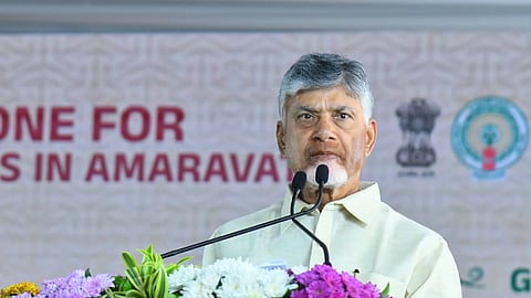 A close-up photograph shows N. Chandrababu Naidu, the former Chief Minister of Andhra Pradesh, speaking at a lectern adorned with a large floral arrangement of white, purple, and yellow flowers. He is wearing a light-colored, possibly cream or pale yellow, Indian garment. Behind him, a banner is partially visible with text related to 'AMARAVATI' and 'Chief Minister.'