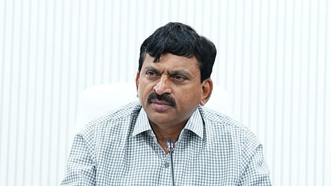 A portrait photo of a man seated at a desk, looking forward with a serious expression. He has dark hair, a mustache, and is wearing a light-colored, checkered button-down shirt. In the background, centered above him on a white, vertically-striped wall, is the circular Emblem of the Government of Telangana in gold and blue. There is a microphone in front of him on the desk.
