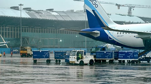 An Indigo airline Airbus A320 aircraft is parked on a wet tarmac at Chennai International Airport. A ground support vehicle (a pushback tug) is towing a train of blue cargo containers labeled "IndiGo Ground Force" near the tail section of the airplane. Construction is visible in the background.