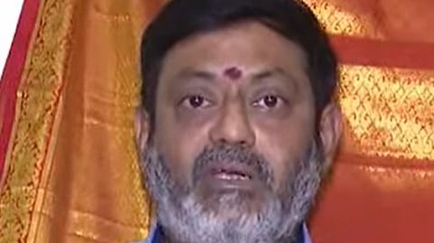 A middle-aged man with a short, salt-and-pepper beard is looking directly at the camera. He is wearing a light blue collared shirt with a small emblem on the chest. He has a red tilak mark on his forehead, and a backdrop of orange and red fabric is visible behind him.