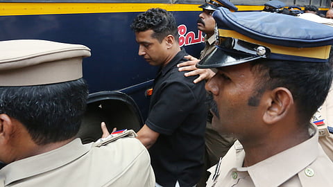 A man in a black shirt is surrounded and escorted by uniformed police officers as he is taken towards a police vehicle outside a court complex.