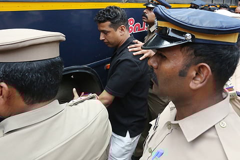 A man in a black shirt is surrounded and escorted by uniformed police officers as he is taken towards a police vehicle outside a court complex.