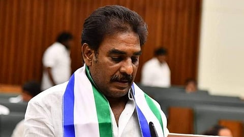 Pinnelli Ramakrishna Reddy is standing at a podium in what appears to be a legislative assembly or parliament. He is wearing a white shirt and a scarf draped around his neck that has the colors and insignia of the YSR Congress Party (YSRCP) – blue, white, and green stripes. He has a mustache and is looking down at a small white folder or tablet on the podium as he speaks. The background is slightly blurred, showing other individuals seated or standing in the assembly hall.