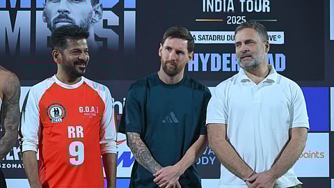Three men standing side-by-side in front of a large black backdrop with white and orange text. From left to right: Revanth Reddy wearing an orange and white long-sleeved soccer jersey with the number 9 and initials "RR" on the chest, Lionel Messi wearing a dark teal short-sleeved shirt, and Rahul Gandhi wearing a white polo shirt. The backdrop text includes "INDIA TOUR 2025" and "HYDERABAD."