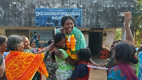 A joyful outdoor scene in front of a Gram Panchayat (village council) office in Amaragiri, Telangana, India. In the center, a tall woman wearing a teal saree and a vibrant orange and yellow marigold garland laughs heartily as she is embraced by two other women.
Around them, several other women in colorful sarees celebrate with broad smiles; one woman on the right has her arm raised in the air in a gesture of victory or cheer. The building in the background features a blue sign with Telugu script that translates to "Gram Panchayat Office, Amaragiri."