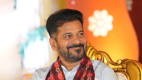 A medium shot of Revanth Reddy, the Chief Minister of Telangana, sitting in an ornate gold-framed chair and smiling. He has a beard and is wearing a light blue button-down shirt with a red and maroon patterned traditional scarf (kanduva) draped around his neck. He is looking off-camera to the right. The background is softly blurred, showing warm orange and yellow lighting with a white floral decoration.