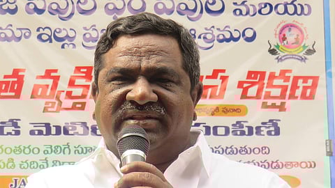 A medium-to-close-up shot of Gade Innaiah, a middle-aged man with dark hair and a mustache, wearing a crisp white button-down shirt. He is holding a microphone with both hands close to his face, appearing to be giving a speech or presentation.
Behind him is a large banner with text in Telugu script. The banner features blue and red lettering on a white background, with a small circular logo on the right side depicting a stylized figure. The man has a serious yet focused expression, and a pen is visible clipped to his shirt pocket. The lighting is bright and even, suggesting an indoor or shaded outdoor event.
