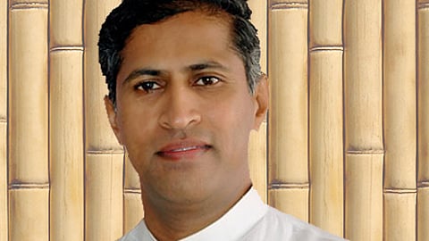 A portrait of Manthena Satyanarayana Raju, a well-known Indian naturopath, from the chest up. He is wearing a traditional white mandarin-collar shirt and has a calm expression. The background consists of vertical light-brown bamboo poles.