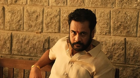 A medium shot of a man with a beard and mustache, wearing a light-colored button-down shirt and gold jewelry, including rings and a bracelet. He is sitting on a wooden bench against a stone wall, looking directly at the camera with a serious expression and holding a phone in his hand.